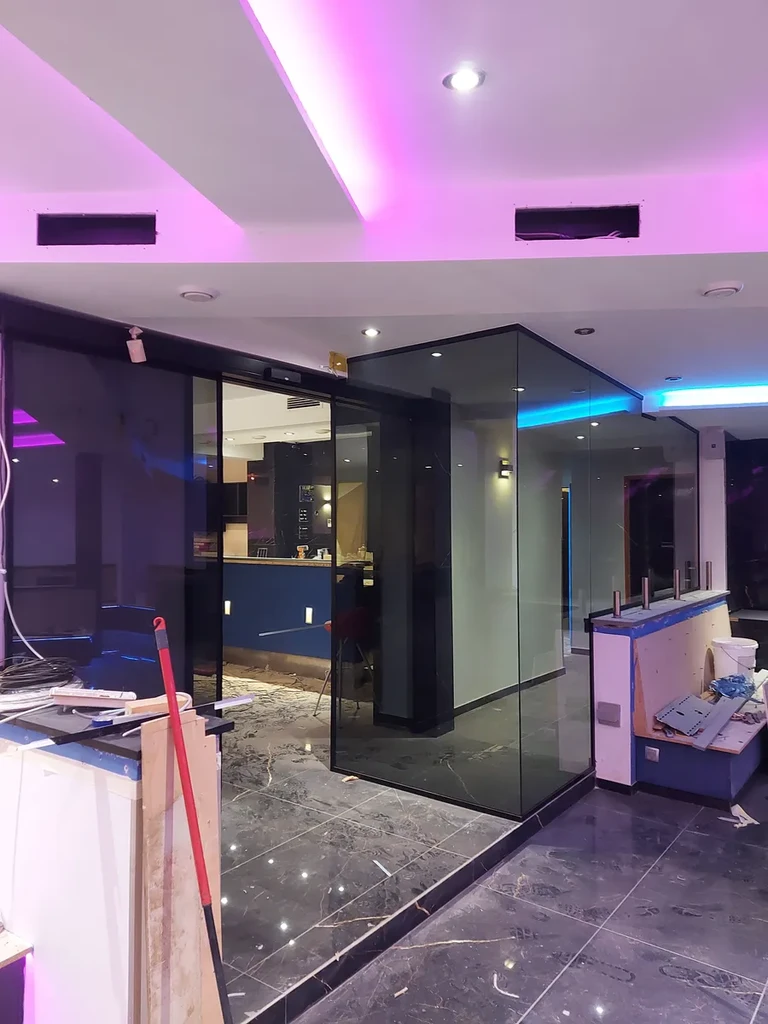 Predelna stena iz temnega kaljenega stekla z reflektnim premazom — učinek temnega ogledala. Steklo po meri, črni profili, kotni spoj 45°. Lounge & Caffe Bar WUDU, Center, Ljubljana